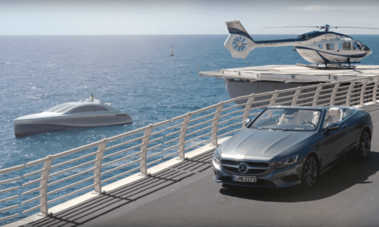 A luxury scene features a gray convertible Mercedes-Benz driving on a road parallel to the ocean. Nearby, a sleek white yacht sails on the water, and a blue and white helicopter rests on a heliport overlooking the sea.