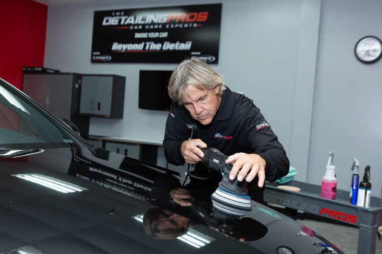 A person in a workshop is polishing a black car with a power buffer. The workshop features a sign that reads, "The Detailing Pros" along with other equipment and detailing supplies from the Revivify Coatings Gallery in the background.