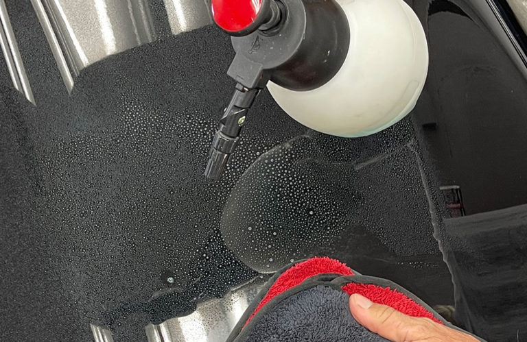 A hand holds a red and black microfiber cloth, wiping a wet black surface. A spray bottle with a red nozzle and white container rests nearby while water droplets glisten around it. The background, reminiscent of the Revivify Coatings Gallery, showcases reflections and shiny areas.