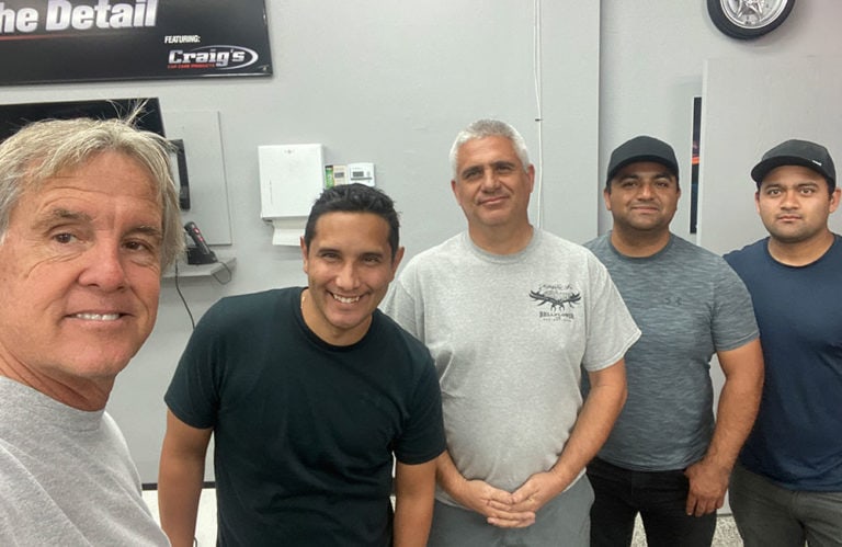 Five men are standing indoors and smiling at the camera in what appears to be the Revivify Coatings Gallery. The man on the left is taking a selfie, and the other four are standing in a line behind him. They are casually dressed, with a sign on the wall behind them reading "The Detail".