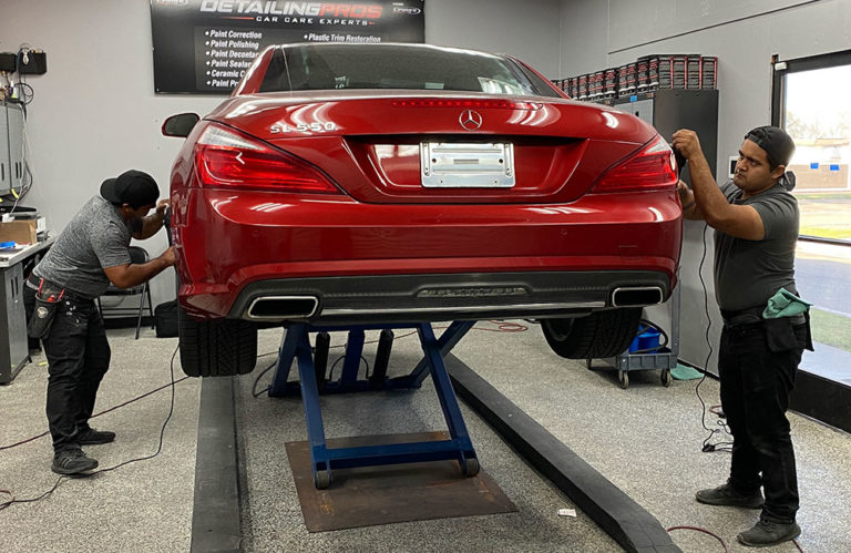 Two technicians from Revivify Coatings Gallery detail a red Mercedes-Benz SL550, elevated on a blue hydraulic lift inside an auto garage. They are working on the car's rear-right and left sides, using tools. The car's rear license plate frame is empty, and a large garage door is visible in the background.