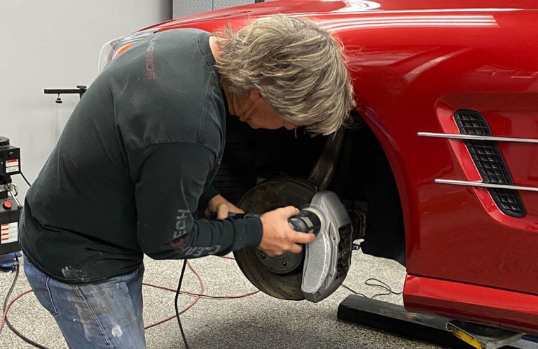 A person with grey hair, wearing a black long-sleeve shirt and jeans, is working on the brake system of a red car inside Revivify Coatings' garage. The car's front wheel is removed, exposing the brake disc and components, creating a scene akin to an automotive gallery.