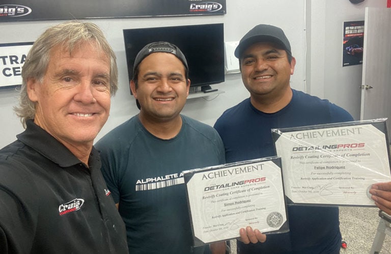 Three men are standing indoors, smiling at the camera. The man on the left is wearing a gray Craig's shirt, the middle man is in a dark Alphalete Athletics shirt and cap, and the man on the right is in a cap, holding two certificates of achievement for detailing at Revivify Coatings Gallery.