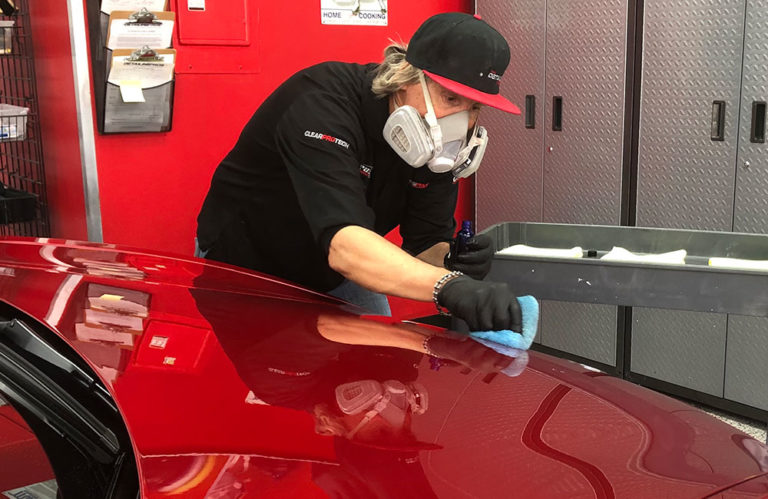 A person wearing a cap, safety goggles, and a respirator mask is polishing the shiny red hood of a car with a blue cloth in the Revivify Coatings Gallery, surrounded by striking red and gray walls.