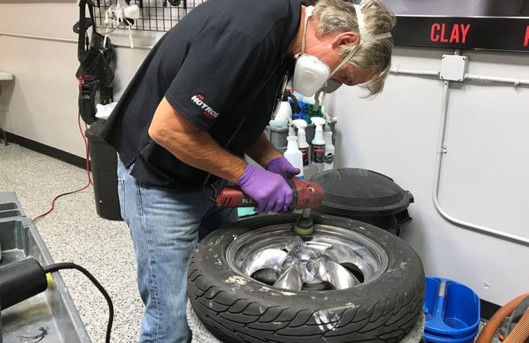 A person wearing protective goggles, mask, and gloves uses a power tool to polish a car wheel in the workshop at Revivify Coatings Gallery. Shelves stocked with various tools and car care products are visible in the background.