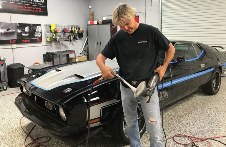 A person with blonde hair is using a power buffer on a black car in Revivify Coatings Gallery. The car boasts blue and white stripes and a Mustang emblem. Dressed in a black T-shirt and ripped jeans, the individual works meticulously amidst various tools and equipment adorning the garage's walls and shelves.