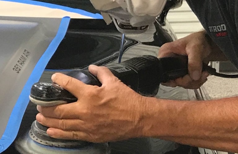 A person using a handheld power tool to polish a car's surface in the Revivify Coatings Gallery. The tool is near a blue tape-lined edge on the car, and the person's hands are firmly gripping the device. Part of the car's exterior is visible, reflecting the surroundings.