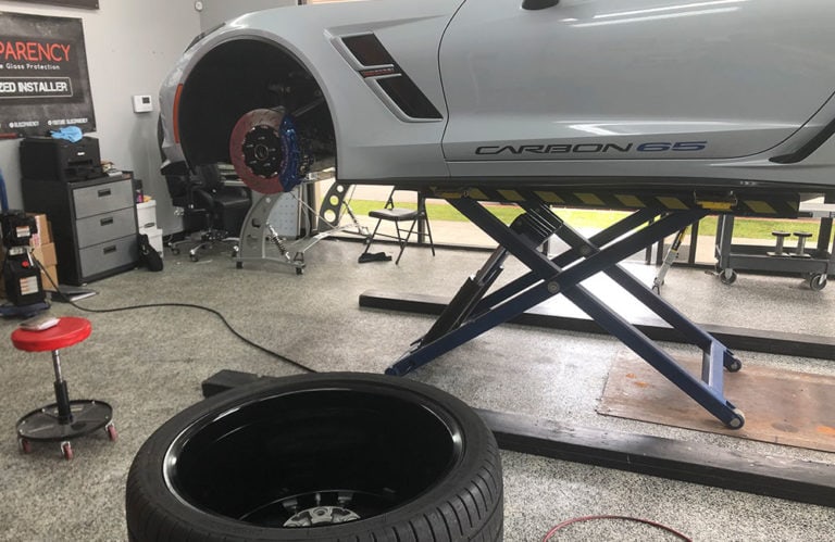 A white car elevated on a hydraulic lift in an auto repair shop, with its front tire removed revealing the brake rotor. A dismounted tire lies in front on the floor. Various tools and equipment are visible around the shop, including a rolling stool and a workbench, showcasing the meticulous environment of Revivify Coatings Gallery.