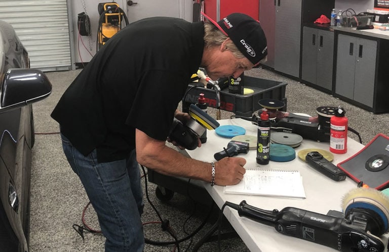 A man wearing a black cap and shirt is standing and writing on a notebook placed on a table in the Revivify Coatings Gallery workshop. The table is covered with car polishing equipment, including polishers, pads, and bottles. A black car is visible on the left. The workshop has a metallic garage door.