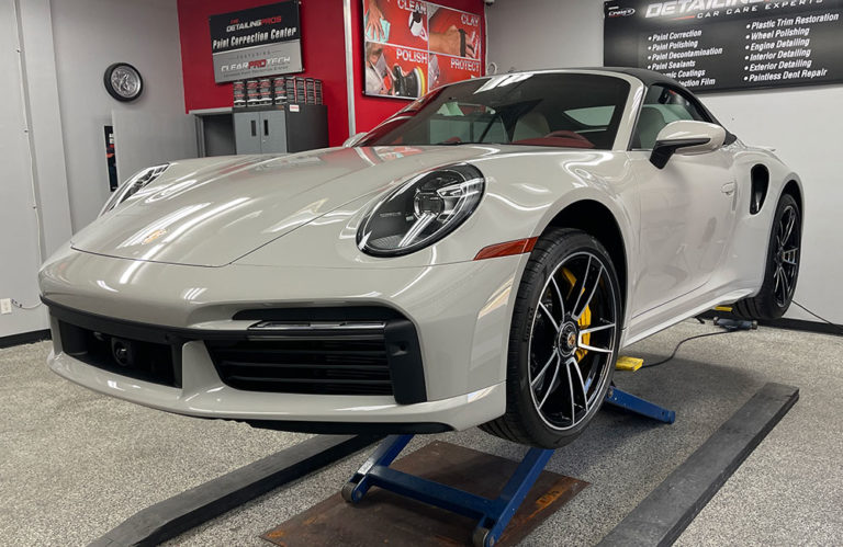 A sleek grey sports car is raised on a hydraulic lift in the Revivify Coatings Gallery indoor detailing center. The car boasts a distinctive front design and black alloy wheels with yellow brake calipers. Red and grey walls, detailing equipment, and various automotive signage serve as the backdrop.