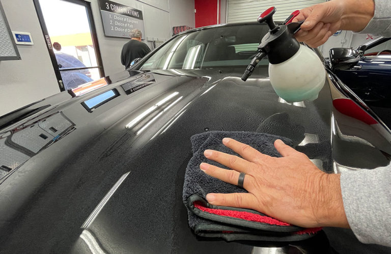 In the Revivify Coatings gallery, a person cleans the hood of a black car inside a garage using a spray bottle and a microfiber towel. The reflection of lights on the car's surface is visible, and other cars and work equipment can be seen in the background.