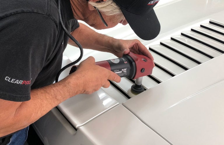 A person wearing a black cap and shirt uses a red electric buffer to revivify the surface of a white car. The car has black vent-like features on its hood, and the person is focusing on the area next to them in what appears to be the Revivify Coatings Gallery.