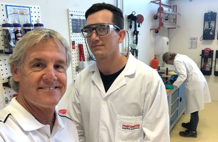 Two men in white lab coats smile in a workshop. The man on the right wears safety goggles and has "Menzerna" printed on his coat. Shelves with tools and equipment line the wall behind them, reminiscent of a Revivify Coatings Gallery, while a third person works at a counter in the background.