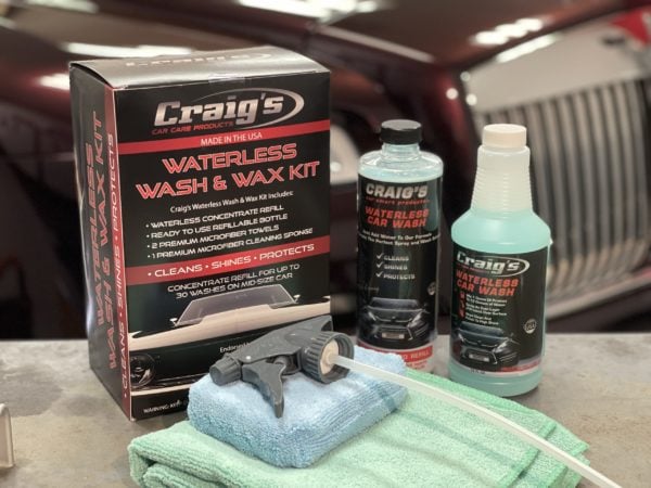 A box labeled "Craig’s Waterless Wash & Wax Alternative for Drought" is displayed on a table alongside a spray nozzle, a green microfiber cloth, a bottle of cleaner, and a bottle of wash concentrate. Ideal for drought conditions, this wash & wax alternative keeps your maroon car gleaming without water waste.