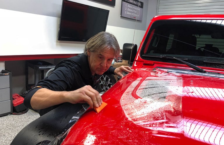 In the Revivify Coatings Gallery, a man is applying a protective film to the hood of a bright red vehicle in a workspace. He uses a small, orange tool to smooth out the film. A black monitor and shelves with various tools can be seen in the background.