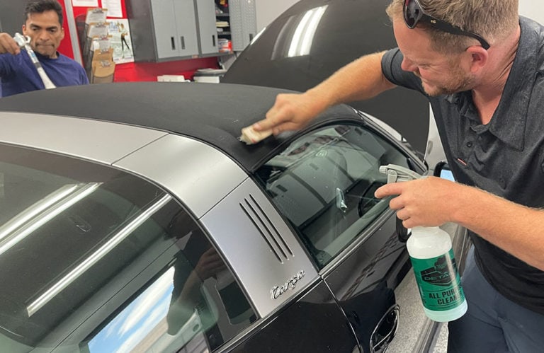 A man is cleaning the convertible roof of a silver car using a small brush while holding a bottle of cleaner from Revivify Coatings. Another man in the background, near shelves with various items in the gallery, holds a small towel. The car’s hood is open, and the setting appears to be a garage.