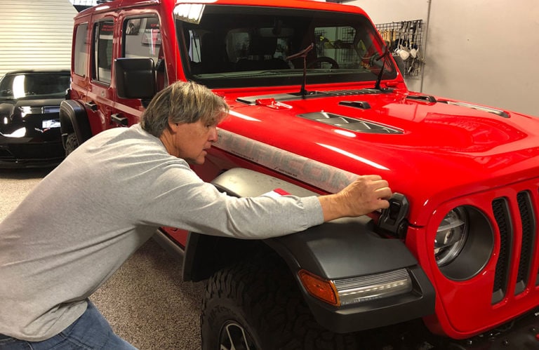 A person with gray hair, wearing a gray long-sleeve shirt, is adjusting or inspecting the front light of a red Jeep vehicle in the Revivify Coatings Gallery garage. The background includes another dark vehicle, tools, and equipment hanging on the wall.