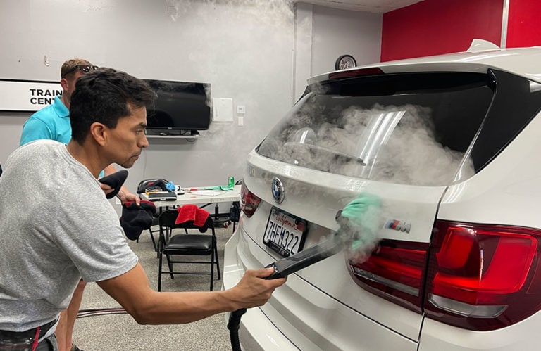 A person uses a steam cleaning device on the back of a white SUV, focusing on the rear area with a license plate reading "7AH5222." Another person, partially obscured, stands nearby. The room, part of the Revivify Coatings Gallery training center, contains various cleaning supplies.