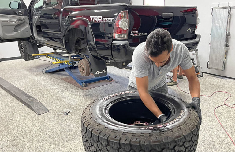 A mechanic in a grey t-shirt is working on a car tire in a garage. In the background, a black Toyota TRD Sport truck is elevated on a hydraulic lift with its rear wheel removed. The mechanic appears to be handling the tire near the wheel of the vehicle, perhaps preparing it for installation at Revivify Coatings Gallery.