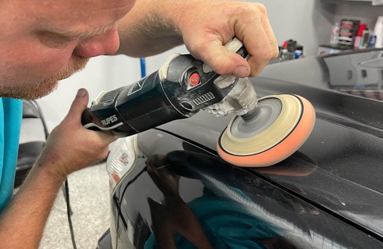 In the Revivify Coatings workshop, a person uses a polishing machine with a soft pad to buff the surface of a black car, focusing on a small section of the rear quarter panel. Various tools and products can be seen in the background, adding to this vibrant gallery of automotive care.