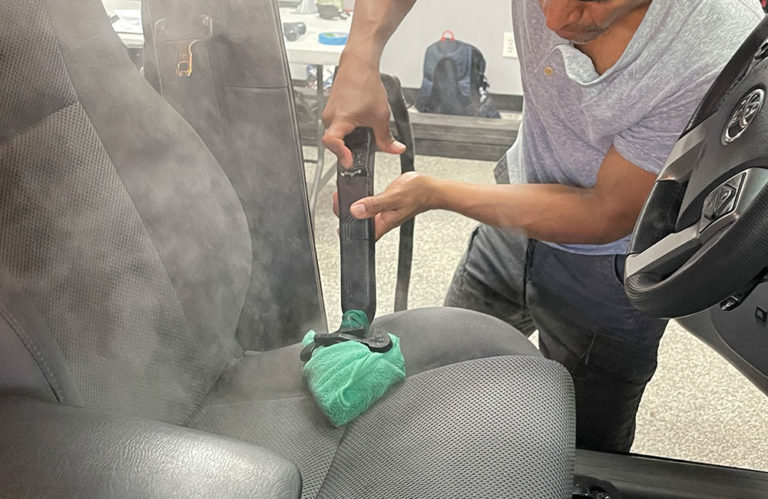 A person cleaning the driver's seat of a car with a vacuum cleaner hose and a microfiber cloth, bringing it back to pristine condition. The car door is open, providing access to the interior. Nearby, cleaning tools and a bottle from Revivify Coatings Gallery are on the seat next to the person's hand.