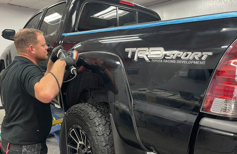 A person is polishing the rear fender of a black pickup truck with a "TRD Sport" decal on its side in the Revivify Coatings Gallery. They are using a polishing machine in a workshop setting, with tools and equipment visible in the background. The truck's paint is glossy and reflective.