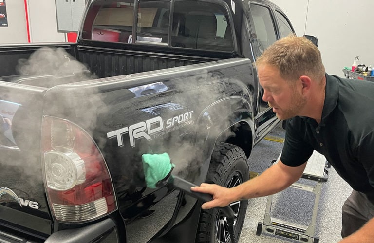 In the Revivify Coatings Gallery, a man uses a steam cleaner to revitalize the black tailgate of a pickup truck marked "TRD Sport" and "V6." He holds a green microfiber cloth at the end of the steam cleaner. The setting appears to be a garage or workshop.