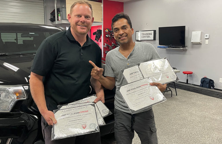 Two smiling individuals stand side by side, holding several framed certificates of achievement at the Revivify Coatings Gallery. They are in a well-lit room with a black vehicle, tools hanging on the wall, and a TV in the background. One person points towards the certificates.