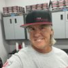 A smiling person wearing a Snap Back Cap and a gray shirt stands in what appears to be an auto detailing or repair shop. Shelves with car care products and metal cabinets are in the background.