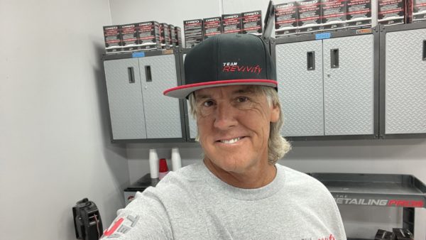 A smiling person wearing a Snap Back Cap and a gray shirt stands in what appears to be an auto detailing or repair shop. Shelves with car care products and metal cabinets are in the background.