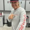 A man stands in a garage, smiling and pointing at his Long Sleeve T-Shirt with "REVVify" written on the sleeve and chest. He wears a black cap with the same logo. Shelves with car products are visible in the background, highlighting his pride in representing REVVify from cap to T-shirt.