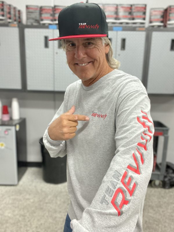 A man stands in a garage, smiling and pointing at his Long Sleeve T-Shirt with "REVVify" written on the sleeve and chest. He wears a black cap with the same logo. Shelves with car products are visible in the background, highlighting his pride in representing REVVify from cap to T-shirt.