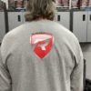 A person with long hair, wearing a black cap and a gray Long Sleeve T-Shirt featuring a red and white logo of a large "R" inside a shield on the back, stands facing a row of lockers. The setting appears to be a garage or workshop with metal cabinets and various items on top.