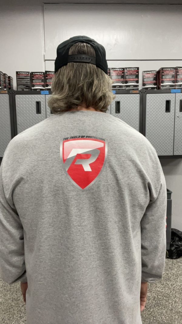 A person with long hair, wearing a black cap and a gray Long Sleeve T-Shirt featuring a red and white logo of a large "R" inside a shield on the back, stands facing a row of lockers. The setting appears to be a garage or workshop with metal cabinets and various items on top.