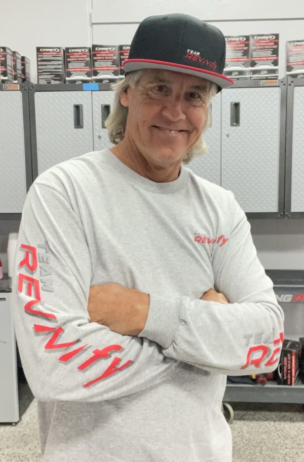 A person with long hair is smiling and standing with arms crossed, wearing a black baseball cap that says "Team Revvvify" and dressed in a light gray Long Sleeve T-Shirt featuring the "Team Revvvify" logo in red on both the front and sleeves.