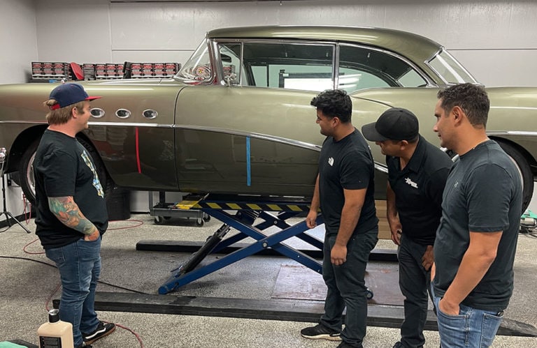 Four men are standing around a classic green car that is elevated on a hydraulic lift in the well-lit Revivify Coatings Gallery. They appear to be discussing something related to the car, with tools and equipment visible in the background.