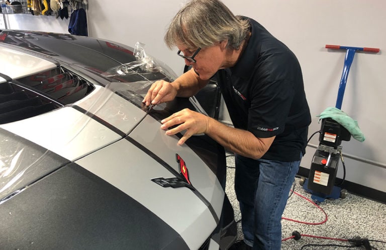 A man with gray hair and glasses is carefully applying a dark stripe decal to the rear of a silver car with a screwdriver in hand. Inside the garage, which resembles the Revivify Coatings Gallery, tools and equipment are visible in the background.