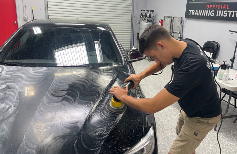 A man is polishing the hood of a black car using a power tool, its surface partially covered in white polishing compound. Focused on his work inside the Revivify Coatings garage, he toils amidst various tools and equipment, with a sign reading "Official Training Insti..." in the background.