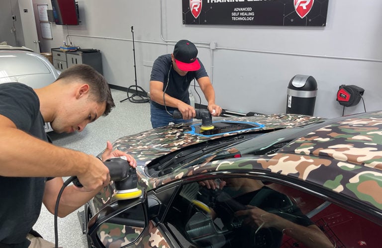 Two men are using polishers on the roof of a camouflage-patterned car in the Revivify Coatings Gallery workshop. Both are focused on their tasks, one wearing a red and black cap. The workshop has various tools and equipment, with a sign on the wall displaying "Training Academy.