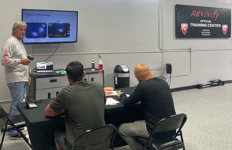 An instructor stands at the front of a classroom, pointing to a screen displaying a presentation about paint damage. Two seated individuals take notes. The room, adorned with a "Revivify Training Center" sign on the wall and various equipment, also highlights the Revivify Coatings Gallery.