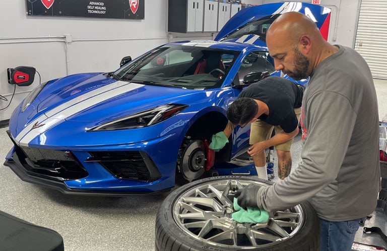 Two men work on a blue sports car in a garage. One is cleaning the brake components with a red cloth while the car is raised on a jack. The other man, under the gleaming lights of Revivify Coatings Gallery, is polishing a car wheel with a green cloth. The garage has a clean, organized appearance with tools in the background.