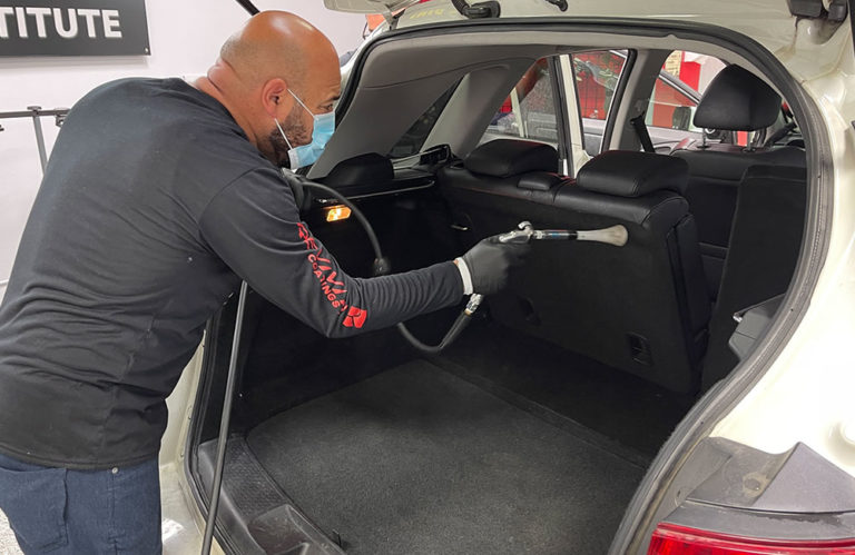 A person is seen cleaning the trunk of a vehicle with a steam cleaner at Revivify Coatings Gallery. They are wearing a black long-sleeved shirt, gloves, and a face mask. The vehicle's rear seats are folded, revealing a spacious trunk. A black and white sign is visible in the background.