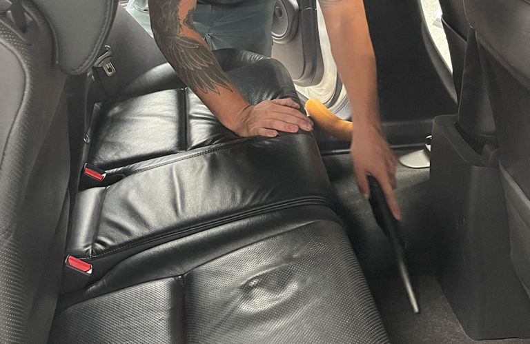 A person with a tattooed arm is vacuuming the back seat of a car with black leather upholstery. The vacuum's nozzle is directed at the car's floor, and the car door is open, allowing light to shine in, highlighting the pristine finish that looks straight out of a Revivify Coatings Gallery.