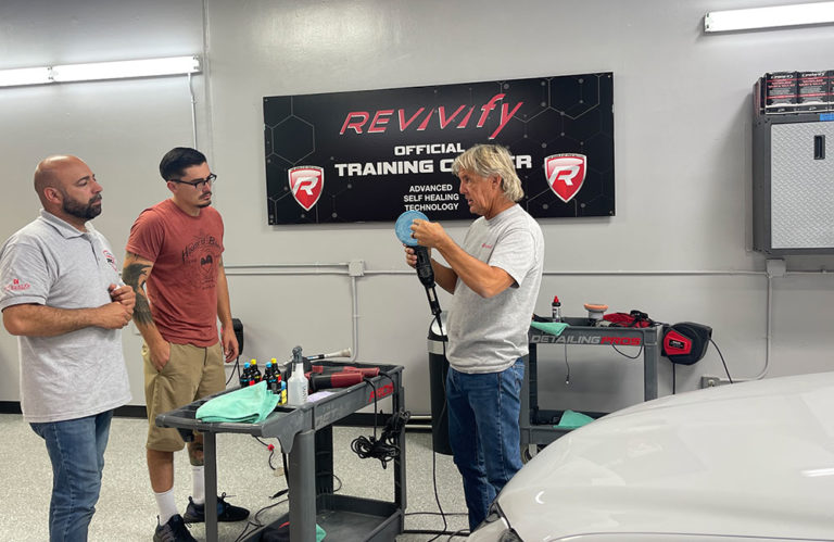 Three men are inside an auto detailing training center. One man is demonstrating the use of a polishing tool while the other two watch attentively. Various detailing supplies are arranged on a nearby cart under a sign that reads "REVIVify Coatings Gallery - Official Training Center.