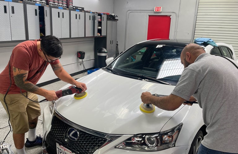 Two people are working on the hood of a white Lexus in a garage. One is using a tool on the left side of the hood, and the other is using a similar tool on the right side. With gray cabinets and a closed red door in the background, it's almost like stepping into an automotive Revivify Coatings Gallery.