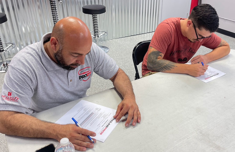 Two men are sitting at a table and signing documents. The man on the left has a bald head and is wearing a gray polo shirt, while the man on the right, participating in the Paint Correction & Vehicle Coating Two Day Training Program With Certification, has short dark hair, glasses, tattoos on his left arm, and is wearing a red t-shirt.