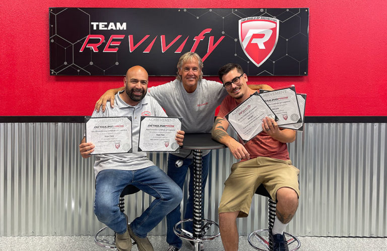 Three men posing together while holding certificates of achievement from the "Paint Correction & Vehicle Coating Two Day Training Program With Certification." They are inside a workshop with a "TEAM REVIVifY" sign in the background. Seated on bar stools against a wall with corrugated metal and a red stripe, they display their accomplishments proudly.