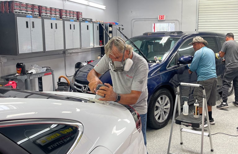 Three men work on cars inside the Revivify Coatings Gallery workshop. In the foreground, a man wearing a mask polishes a white car's surface. Another man in the back cleans a blue car's windows using a cloth. The workshop is equipped with tools and cabinets in the background.