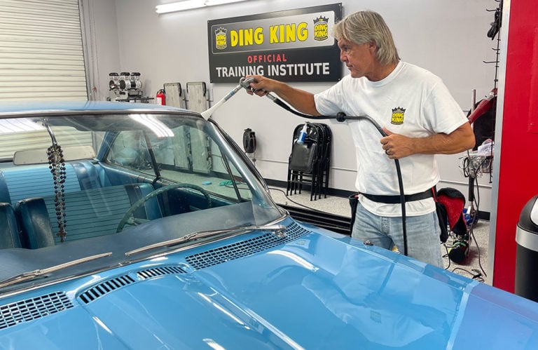 A man in a white T-shirt is steam-cleaning the windshield of a blue car inside the Revivify Coatings Gallery. A sign in the background reads "Ding King Official Training Institute," with chairs and car-related equipment visible in the room.