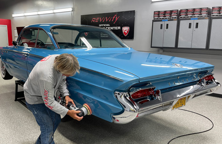 A person polishes the rear fender of a shiny blue vintage car in a garage, elevated on a platform, using a power buffer. The background features cabinets, equipment, and a sign reading "Revivify Coatings Gallery OFFICIAL CENTER.